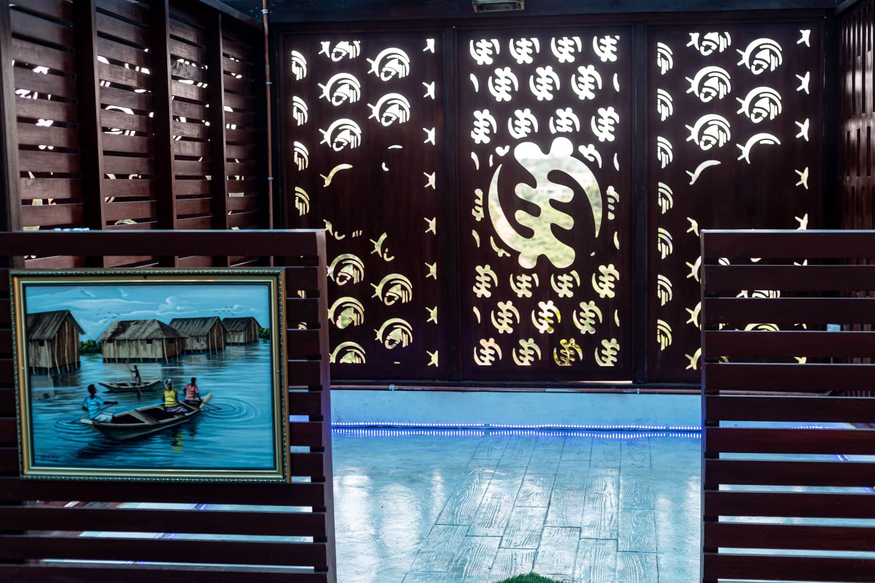A timber screen cut with the Gye Nyame symbol, beside a painting of a stilt village on the water